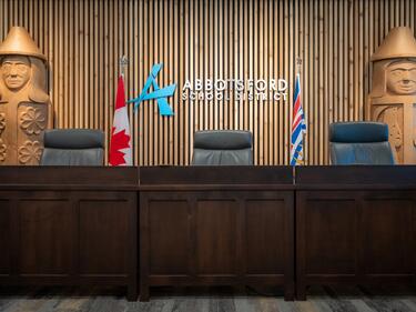 Abbotsford School District Logo on a wood slated wall with the Canadian flag on the left side and the BC flag on the right side of the logo. Desk sits in front and 3 board room chairs.